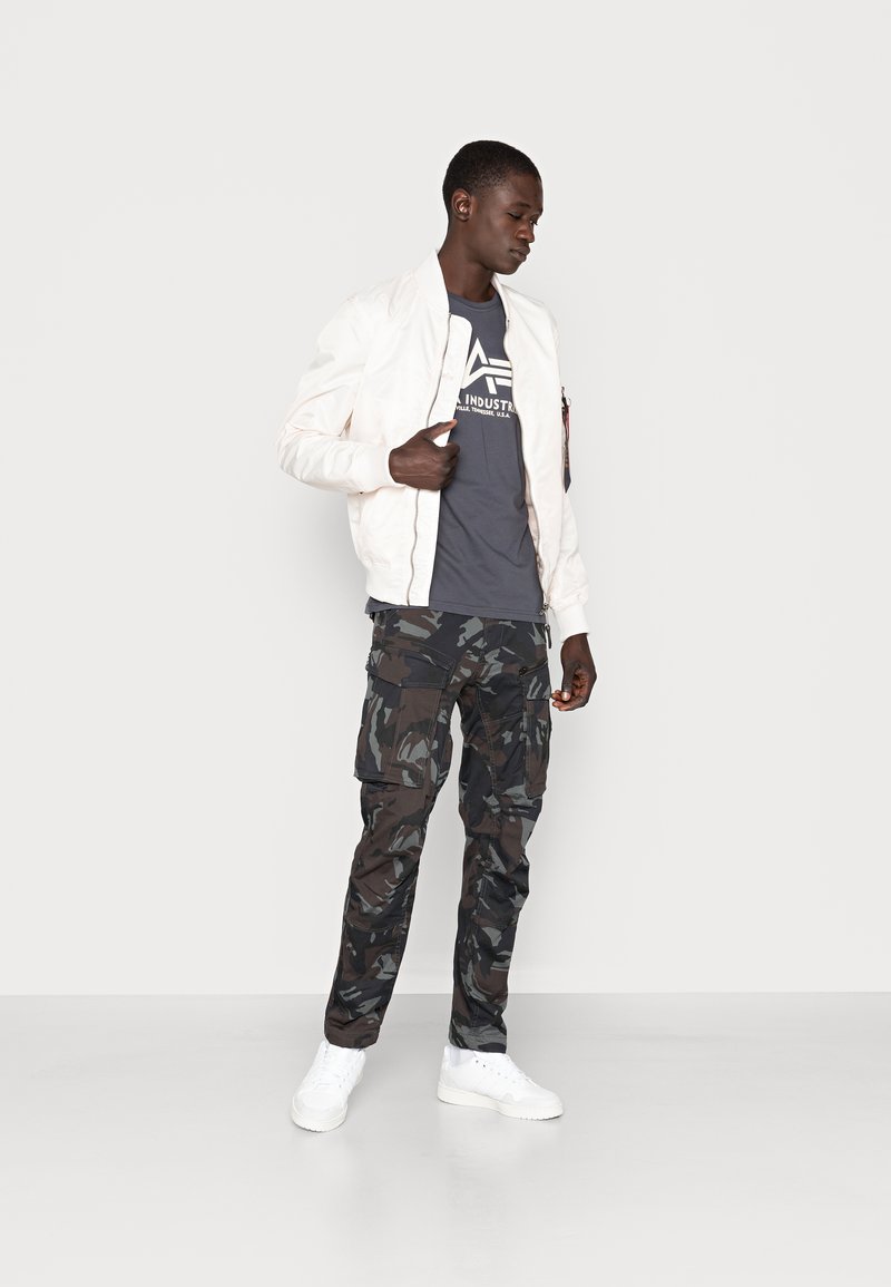 Jachetă bomber albă, tricou grafic gri, pantaloni cargo cu camuflaj și adidași albi; având modele distincte și texturi variate.
