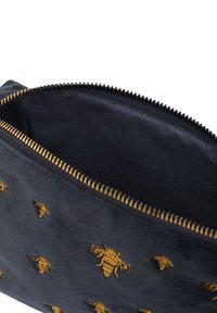 Bolsa de tela negra con un bordado dorado de abejas y una cremallera dorada parcialmente abierta que muestra el forro interior oscuro.