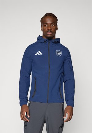 Marineblå zip-jakke med hætte, prydet med hvide Adidas-striber og Arsenal-logo. Indeholder to lynlåslommer på siden og elastiske manchetter.
