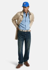 Abrigo trench beige, camisa de botones azul claro, jeans oscuros, zapatos de cuero marrón y una gorra azul con un logotipo. Conjunto minimalista y casual.