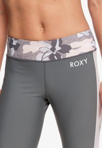 Szare legginsy z pasem w kwiatowy wzór w odcieniach szarości, różu i bieli. Logo marki "ROXY" z boku. Gładka faktura.