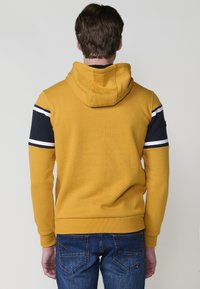 Una persona si trova di spalle rispetto all'osservatore, indossando una felpa con cappuccio gialla con maniche a righe blu e bianche e jeans blu.