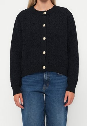 Femme portant un cardigan en tricot texturé noir avec six boutons dorés sur un jean en denim bleu, debout devant un fond uni.