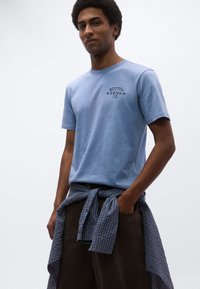 Φωτεινό μπλε βαμβακερό μπλουζάκι με μαύρο λογότυπο στο στήθος. Συνδυασμένο με καφέ παντελόνι και μπλε καρό πουκάμισο δεμένο στη μέση.