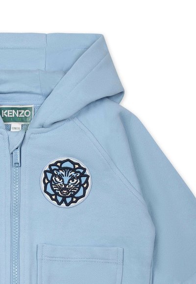 Hoodie zippé bleu clair avec une capuche, une poche avant et un badge de tigre brodé entouré d'un motif floral. Matière douce en coton.