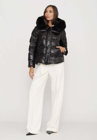 Giacca a piumino nera con finitura lucida e un ampio collo in pelliccia nera. Presenta chiusura con zip e tasche laterali, indossata con pantaloni bianchi.