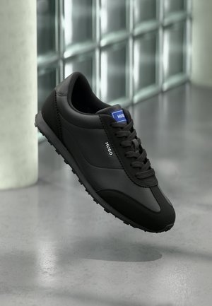 Schwarze Sneakers aus Leder mit glatter Textur, ausgestattet mit einem niedrigen Schaft, rundem Zehenbereich und Schnürverschluss vorne. Blaues Markenlabel vorhanden.