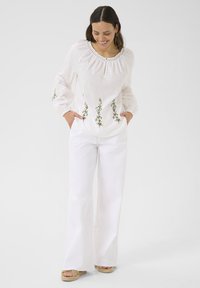 Blusa di lino bianca con scollatura arricciata e ricami floreali in verde e rosa. Abbinata a pantaloni bianchi a gamba larga e sandali platform color beige.
