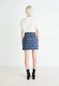 Person with short blonde hair wearing a white T-shirt, patterned blue denim skirt, and black ankle boots standing facing away.