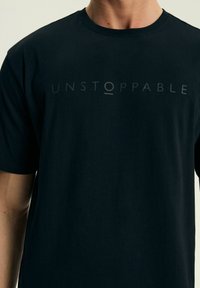 Czarny t-shirt z krótkim rękawem wykonany z miękkiego materiału, z napisem "UNSTOPPABLE" w matowym, kontrastowym druku na przodzie.