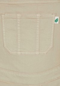 Poche arrière en denim beige avec une double couture verticale et une petite étiquette blanche présentant une feuille verte sur le côté droit.