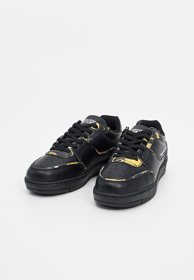 Zapatillas negras con acabado en cuero suave y acentos amarillos, caja de dedos perforada y cordones negros planos. Suela de goma, diseño de corte bajo.