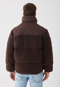 Brun pufferjakke med en fluffy teksturert fleece-kropp og glatt overdel, som har høy krage og ribbede mansjetter.
