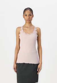 Hellrosa, gerippter Tanktop mit Spitzenbesatz entlang des Ausschnitts und der Armlöcher. Wellenkante am Saum. Getragen mit einem dunkelgrauen Rock.