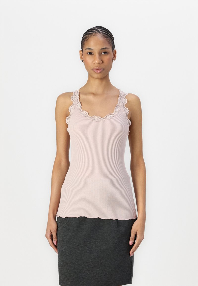 Hellrosa, gerippter Tanktop mit Spitzenbesatz entlang des Ausschnitts und der Armlöcher. Wellenkante am Saum. Getragen mit einem dunkelgrauen Rock.