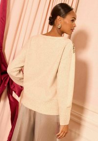 Ženska z bunom, oblečena v krem pulover s bleščicami na rami in svetlo rjavim krilom, stoji ob rožnatih in burgundskih zavesah.