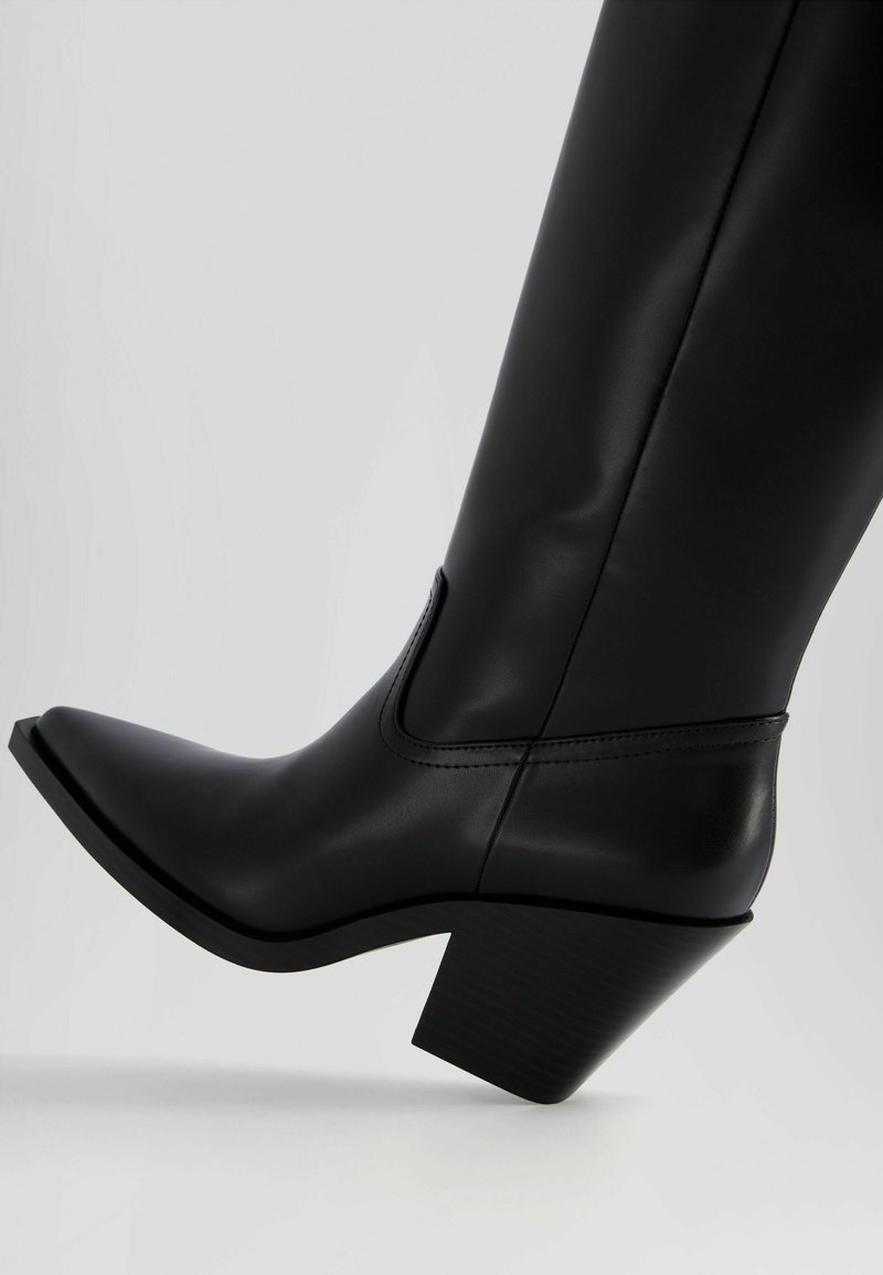 Botte de cheville noire en matériau synthétique lisse, avec un bout pointu, un talon incliné et des détails cousus sur le dessus.