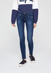 Person som bär mörkblå skinny jeans, en marinblå och vit långärmad sweatshirt och svarta låga sneakers med vita sula.