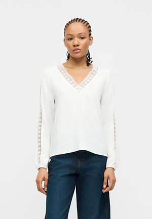 Γυναίκα με πλεξούδες που φοράει λευκό μπλουζάκι με μακριά μανίκια, διακοσμημένο με δαντέλα στο V-λαιμό και τα μανίκια, σε συνδυασμό με σκούρο μπλε τζιν.