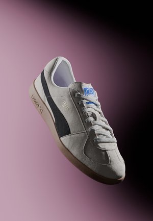 White and black suede Puma sneaker with gum sole, laced up, tilted against a gradient purple to black background.