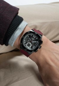 Schwarze und silberne Armbanduhr mit strukturiertem Zifferblatt, ausgestattet mit römischen Ziffern und drei Unterzifferblättern, montiert an einem roten Gummiband.