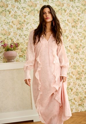 Kobieta o długich ciemnych włosach, ubrana w jasnoróżową sukienkę z falbanami, stojąca w pomieszczeniu z kwiatową tapetą i wazonem z kwiatami za nią.