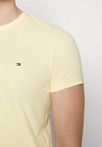 Keltainen puuvillapaita lyhyillä hihoilla, jossa on pieni punavalkosiniset brodeerattu logo rinnan kohdalla. Sileä pinta.