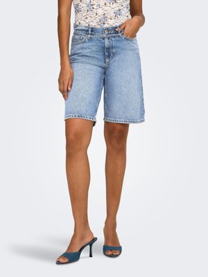 Femme portant un short en jean bleu clair jusqu'aux genoux, un top en dentelle florale et des sandales à talons ouvertes bleues, debout sur un fond neutre.