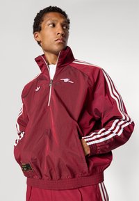 Corta-vento bordô com riscas brancas, gola com fecho de correr e bolso frontal. Apresenta logo no peito e punhos e cintura elásticos. Material leve.