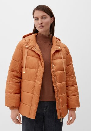 Kobieta w pomarańczowej kurtce puchowej z rozpiętym zamkiem, nosząca brązowy sweter z golfem i ciemne dżinsy, stojąca na tle czystej białej ściany.