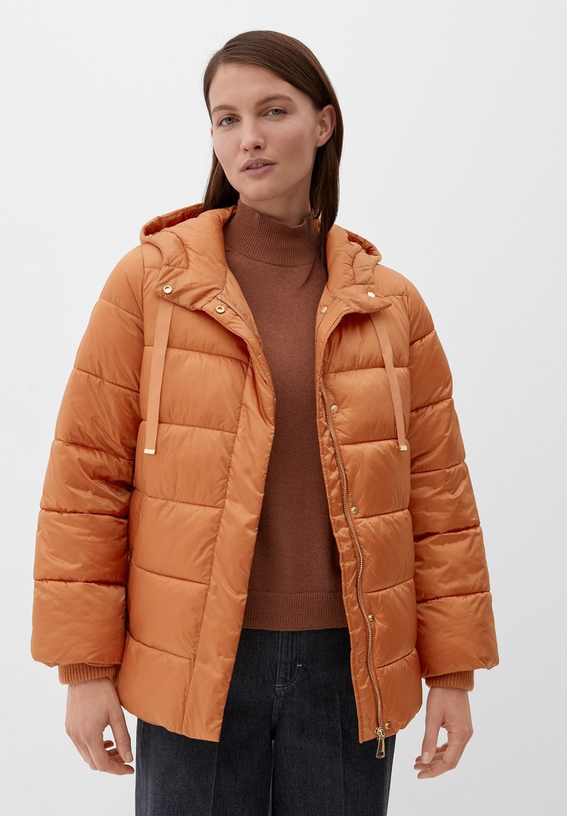 Kobieta w pomarańczowej kurtce puchowej z rozpiętym zamkiem, nosząca brązowy sweter z golfem i ciemne dżinsy, stojąca na tle czystej białej ściany.