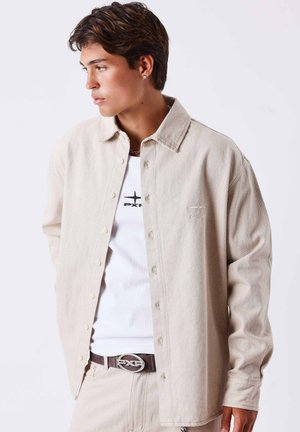 Jeune homme aux cheveux foncés regardant sur le côté, portant une chemise beige boutonnée sur un t-shirt blanc avec le logo PXG et un pantalon beige assorti avec une ceinture.