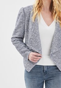 Blazer rayé bleu et blanc en tissu texturé, devant ouvert, manches longues, associé à un haut blanc à col en V et un jean en denim.