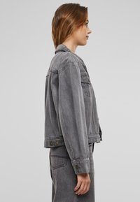 Grå denimjacka med klassisk skärning, med knäppbara ärmslut och framknäppning. Slät textur, minimalistisch design och sidofickor.