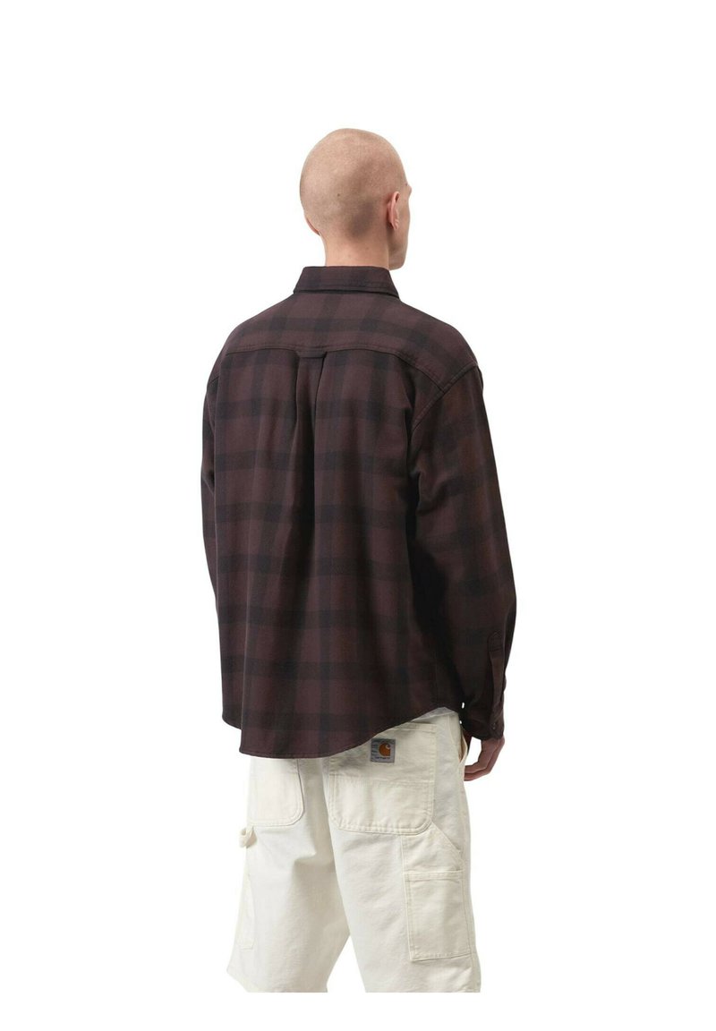 Uomo di spalle che indossa una camicia a quadri scura a maniche lunghe e pantaloni cargo bianchi con un logo del marchio visibile sulla tasca posteriore.
