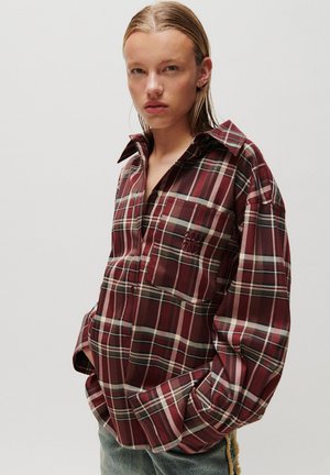 Young person wearing a loose red and green plaid shirt with hands in pockets, standing against a plain light background.