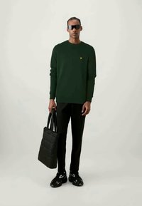 Sweater verde de mangas longas com um pequeno logótipo amarelo, bolsa tote preta acolchoada, calças pretas e sapatilhas pretas desportivas com um design branco.