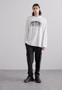 Balta garām piedurknēm krekls ar melnu grafiku, apvienots ar melniem kravas biksēm. Modelis valkā melnus sporta apavus. Vienkāršs fons.