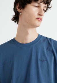 Μπλε βαμβακερό μπλουζάκι με στρογγυλή λαιμόκοψη, κοντά μανίκια και ένα μικρό, σκούρο λογότυπο στο στήθος. Μαλακή υφή και χαλαρή εφαρμογή.