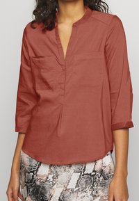 Blouse couleur rouille avec col et deux poches poitrine. Manches trois-quarts et coupe décontractée, fabriquée en tissu léger.