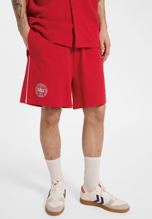 Mężczyzna noszący czerwone sportowe spodenki z białym logo na lewej nodze, białe skarpetki i białe trampki z niebieskimi i czerwonymi detalami.