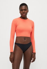 Long-sleeve cropped top in bright coral, textured fabric; paired with black bikini bottoms featuring a minimal design.