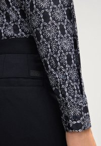 Zwarte shirt met bloemmotief en witte accenten. Bevat een knoopmanchet en contrasterend zwart materiaal van de broek met een label.
