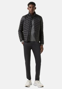 Schwarze, wattierte Jacke mit Chevron-Steppung, hohem Kragen, grauem Sweatshirt, schwarzer Hose und weißen Turnschuhen mit beigefarbenen Akzenten.