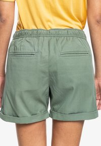 Personne portant un short vert avec ourlets roulés, taille élastique et deux poches passepoilées à l'arrière, associé à un haut jaune.
