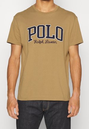 Mężczyzna noszący beżową koszulkę z krótkim rękawem z dużym napisem „POLO Ralph Lauren” w granatowym kolorze oraz czarne dżinsy.