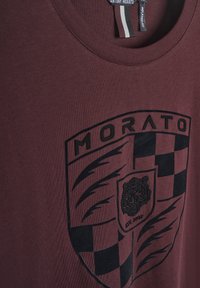 Camiseta de algodón burdeos con un gráfico negro de un escudo que contiene la palabra "MORATO" y un tigre, con acentos de tablero de ajedrez debajo.