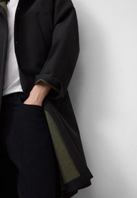 Cappotto nero con una vestibilità ampia, caratterizzato da una fodera interna verde, una tasca sul petto e polsini arrotolati. Indossato sopra una camicia bianca.