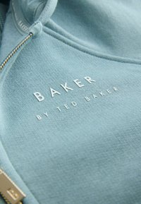 Detailné zábery svetlomodrej látky s bielym logom "BAKER BY TED BAKER" a kovovým zipsom s vyrytým písmenom "B" na mikine.