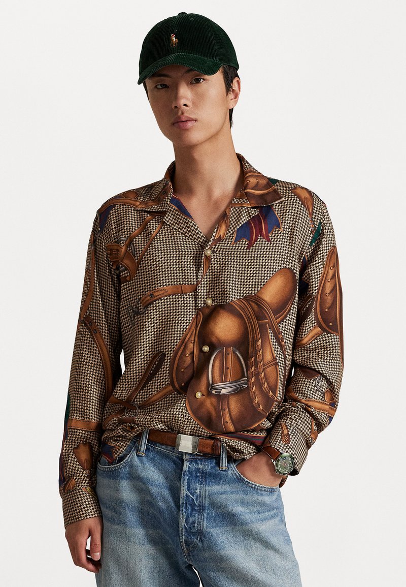 Camisa de manga larga con un patrón de pata de gallo, ilustraciones de sillas de montar y equipo de equitación en tonos tierra. Combinada con jeans de color azul claro.